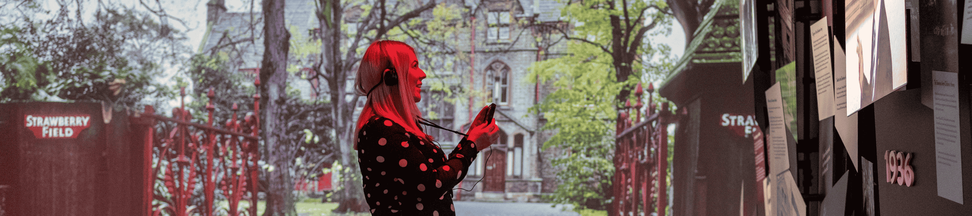 A woman stood in the Strawberry Field exhibition with a headset on. Behind her is a projection of the old Strawberry Field mansion.