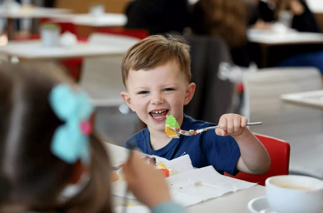 Child eating moutful of food from a spoon while smiling in the bright cafe