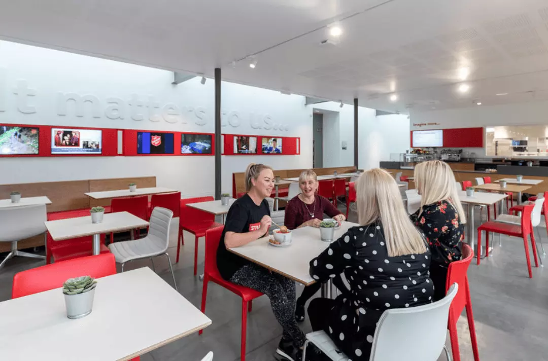 An indoor cafe with huge windows in the sunlight with white tables and red chairs. People are sat eating on this tables and smiling.