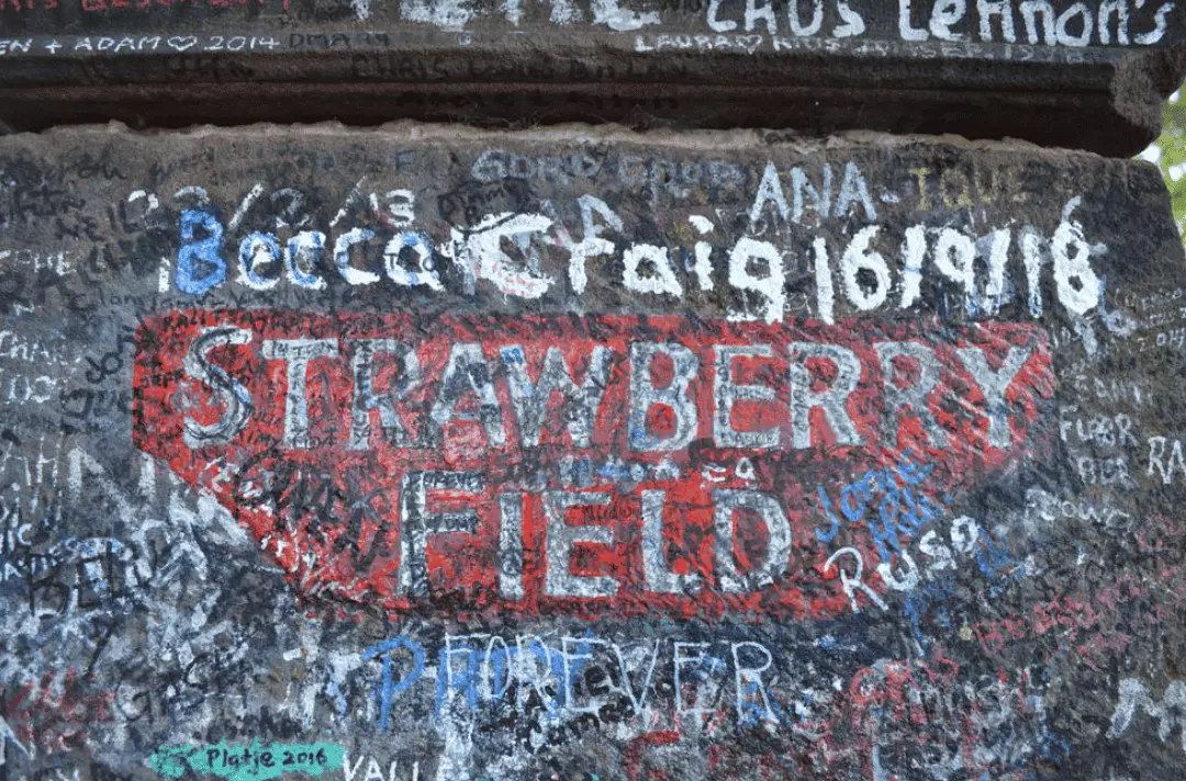Graffiti-covered wall featuring 'Strawberry Field' painted in red and white, surrounded by various signatures, dates, and colorful messages left by visitors