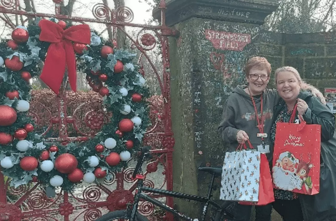 Karen Carter and Sharon Heller stand in front of the Strawberry Field gates with Christmas gift bags and a bike. There is a Christmas wreath on the gate.