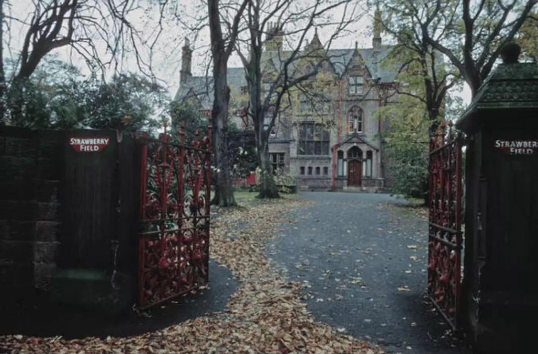 In the foreground is the Strawberry Field red iron gates and gateposts with 'Strawberry Field' painted on them in red and white. In the background is the old gothic mansion that was the children's home.