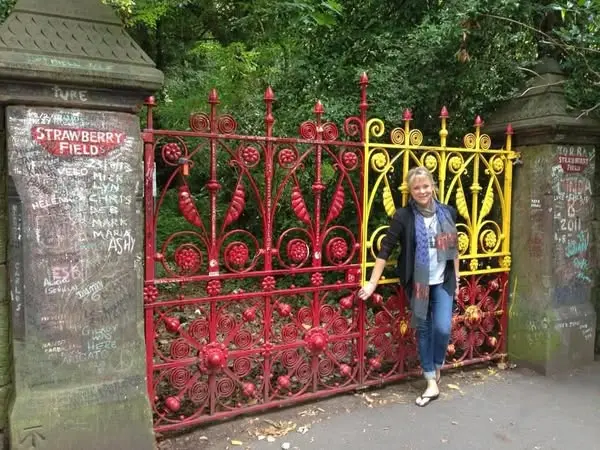 A woman stands in front of the iron gates of Strawberry Field which are painted red with a section painted yellow.