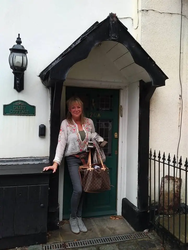 A woman stands in front of a cottage door