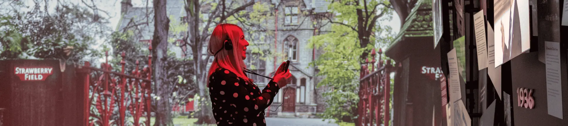 A woman stood in the Strawberry Field exhibition with a headset on. Behind her is a projection of the old Strawberry Field mansion.