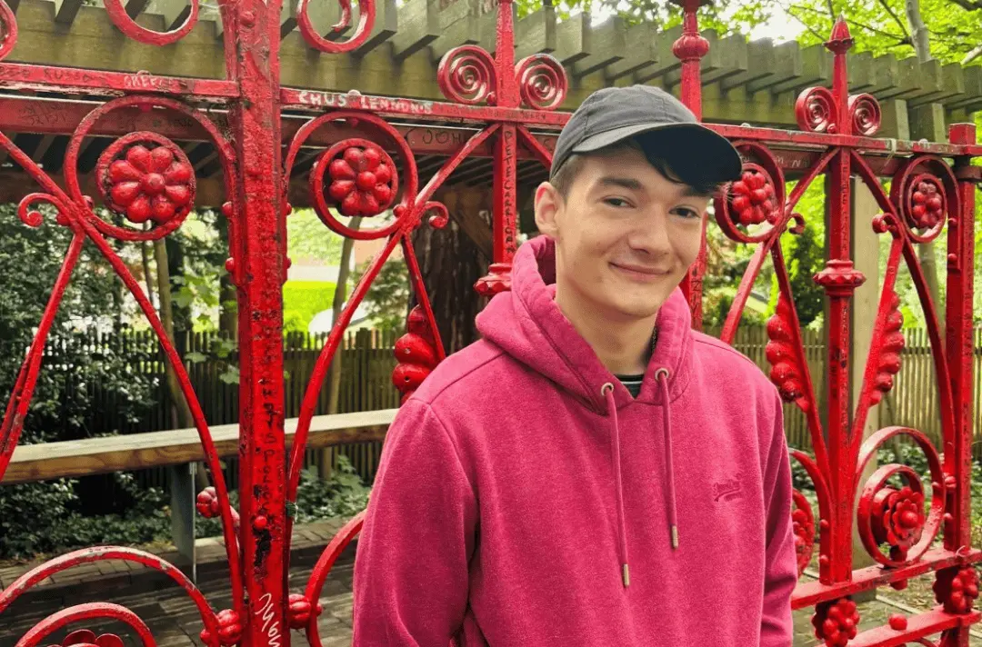 Steff, a male student, stands in front of the red Strawberry Field gates wearing a cap and a red hoodie.