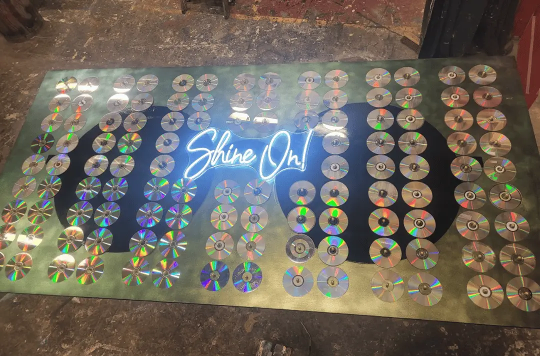Finished artwork, a green board with John Lennon's sunglasses painted on it, a neon light reading 'Shine On!' and rows of CDs.