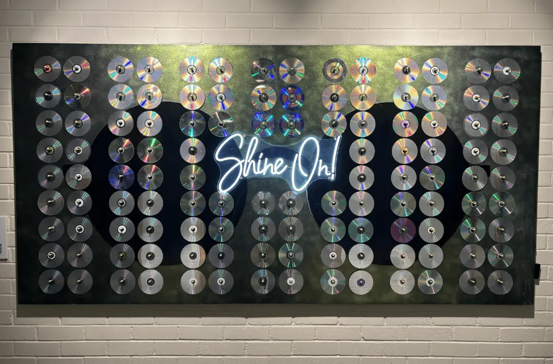 Shine On! by Steff Norman. A large piece of wood painted with green, sparkly paint. John Lennon style round glasses are painted on it and there is a neon sign that says 'Shine On'. There are CDs pegged on to the board.