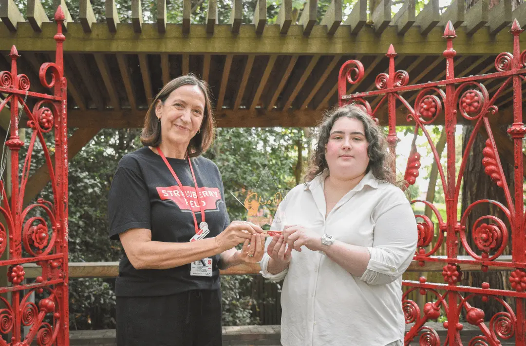 Work coach Becky and Steps to Work trainee Sophie stand between the original Strawberry Field gates holding the 'Shine On!' award