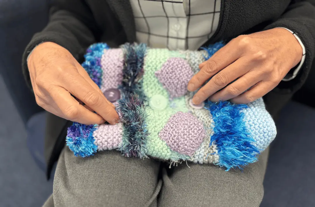 A person holds a knitted fidget muff in their lap
