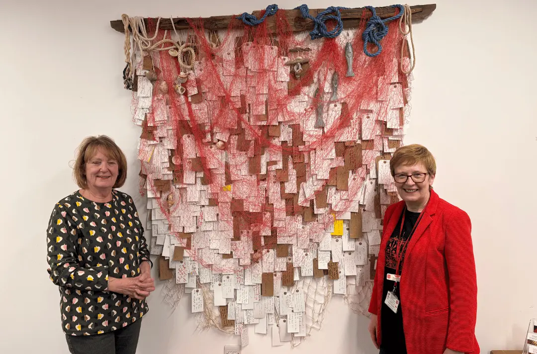 Artist Marilyn Chester with Fresh Expression team member Karen Carter with art piece 'The Catch'. The art work is a piece of drift wood, with hundreds of prayer tags hanging off it and a red fishing net.