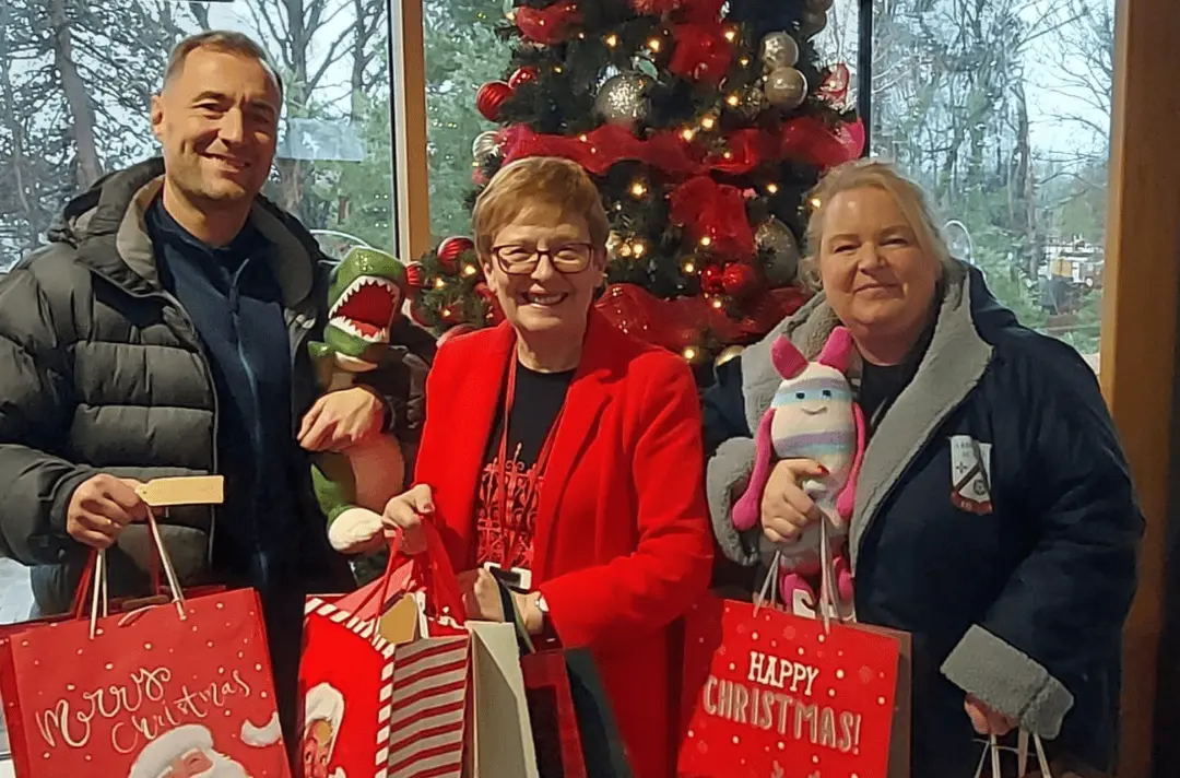 Three people stand in front of a Christmas tree with Christmas gifts